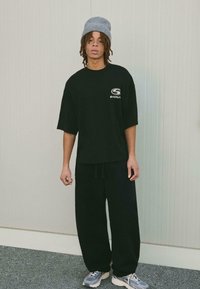 Černé oversized tričko s logem na hrudi, spárované s volnými černými kalhotami. Šedá pletená čepice a šedé běžecké boty dotvářejí celkový vzhled.