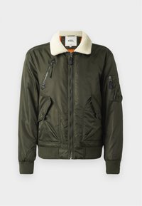 Veste bomber verte olive avec un col en fausse fourrure crème, fermeture éclair à l'avant et plusieurs poches zippées. Manches et bas côtelés. Texture lisse.