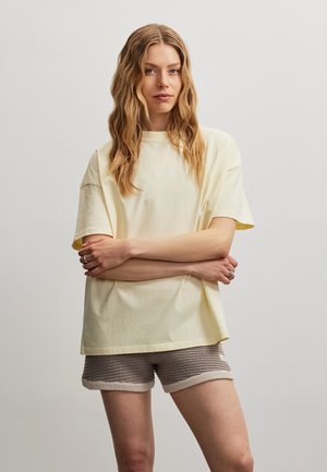 Nuori nainen aaltoilevilla hiuksilla, yllä vaaleankeltainen oversize t-paita ja beige neulotut shortsit, seisoo käsivarret ristissä.