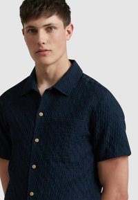 Chemise à manches courtes bleu marine avec un motif texturé, dotée d'un col et de boutons en bois, ainsi qu'une poche sur la poitrine.