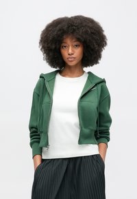 Zelená mikina na zips s vreckami vpredu a kapucňou s nastavovacou šnúrkou, nosená cez biele tričko. Modelka má kučeravé vlasy a má na sebe čiernu plisovanú sukňu.