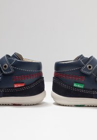 Chaussures en cuir bleu marine avec surpiqûres rouges et accents en daim, dotées de bandes Velcro et d'une semelle en caoutchouc légère. Marque Kickers visible.