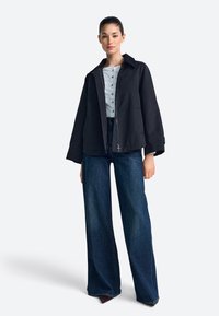 Juoda, lengva striukė su stačiakampio formos siluetu, po ja pilka, mygtukais užsegama marškinėliai, platėjančios tamsios džinsinės kelnės. Lygus audinys ir ryškios linijos.