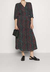 Mörkgrön omlottklänning med röd blommönster, trekvartslånga ärmar, cinchad midja, svängande kjol, ihopparad med svarta slingback-skor och väska.