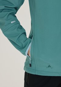 Teal pehmeäkuorijakku, jossa on integroituja taskuja ja heijastava logo; siinä on sileä pinta ja vedenpitävät ominaisuudet.
