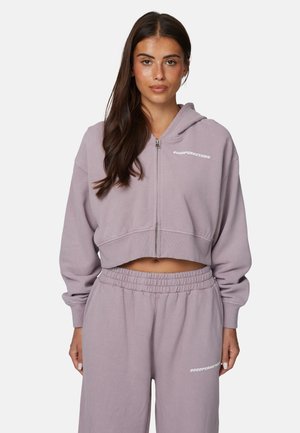 Kobieta ubrana w dopasowaną lawendową krótką bluzę z zamkiem i spodnie dresowe z elastycznym paskiem, na obu elementach biały napis "Good For Nothing".