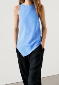 Light blue sleeveless top with round neckline, asymmetrical hem, and relaxed fit, paired with black loose trousers. Smooth fabric texture.