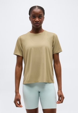 Mujer de pie, de frente, vistiendo una camiseta holgada beige y pantalones cortos de ciclista ajustados de color azul claro frente a un fondo liso.