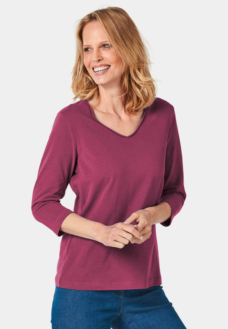 V-Ausschnitt, 3/4-Arm Oberteil aus burgunderfarbenem Baumwollstoff mit glatter Textur. Lässiges Design, kombiniert mit blauen Jeans, Hände vor der Brust gefaltet.