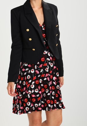 Kvinde iført en sort dobbeltradet blazer med guldknapper over en sort blomsterkjole med røde, hvide og lyserøde blomster.