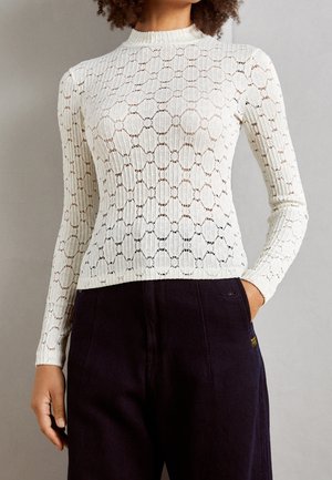 Kobieta ubrana w dopasowaną, białą, koronkową bluzkę z długim rękawem, ozdobioną okrągłymi wzorami i z wysokim kołnierzem, w parze z ciemnymi, szerokimi spodniami.