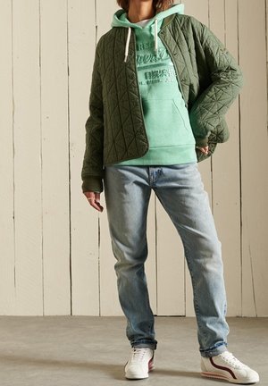 Chaqueta acolchada verde oliva sobre una sudadera con capucha verde claro, combinada con vaqueros azul claro y zapatillas blancas con un toque rojo. Texturas suaves.