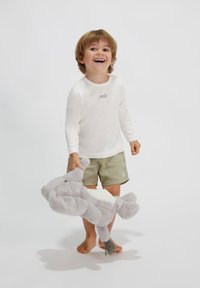 Kind, der ein weißes Langarmshirt mit dem Aufdruck "smile" und grünen Shorts trägt, hält ein graues Plüschelefantenspielzeug. Der Hintergrund ist schlicht.