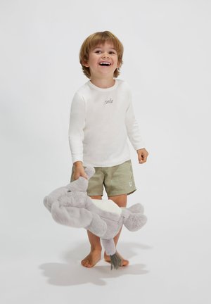 Kind, der ein weißes Langarmshirt mit dem Aufdruck "smile" und grünen Shorts trägt, hält ein graues Plüschelefantenspielzeug. Der Hintergrund ist schlicht.