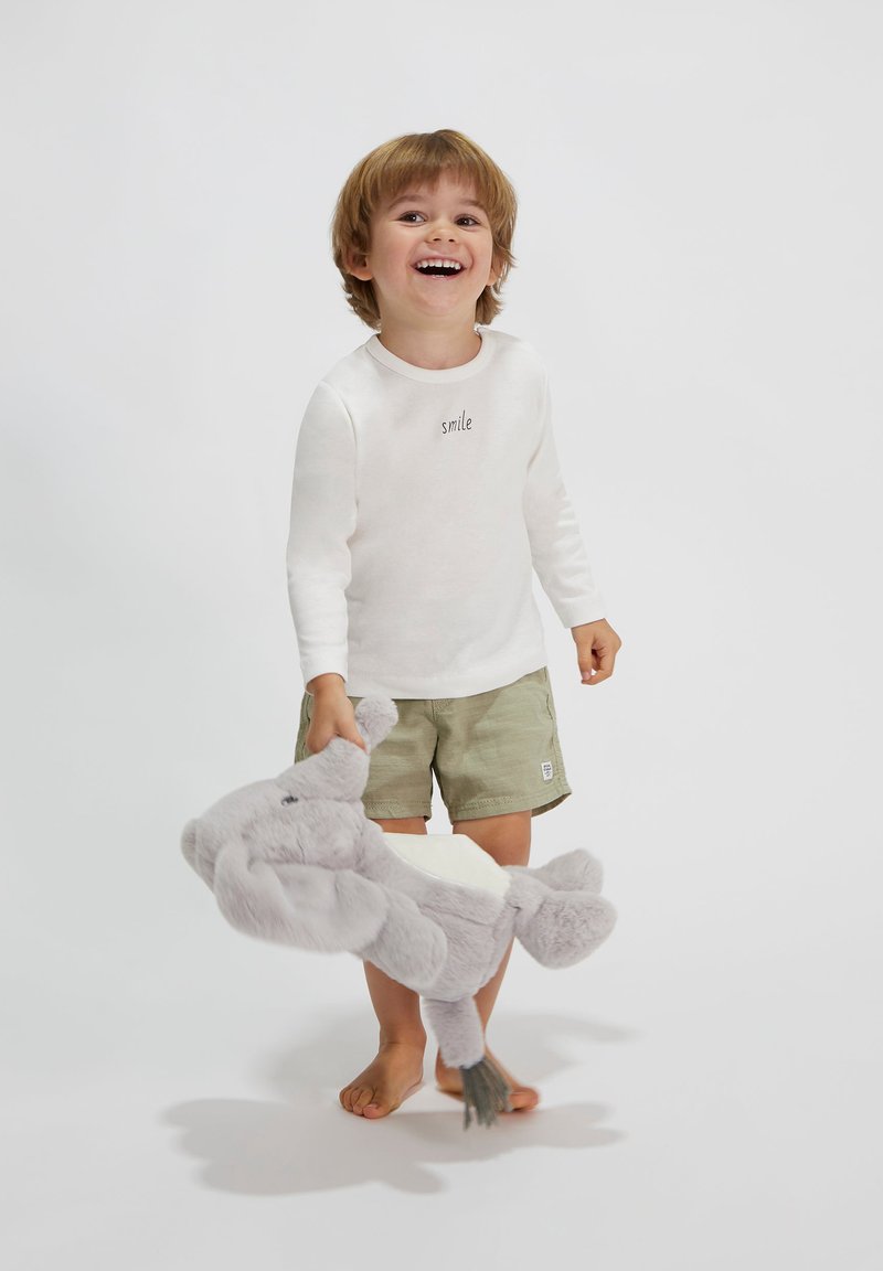 Kind, der ein weißes Langarmshirt mit dem Aufdruck "smile" und grünen Shorts trägt, hält ein graues Plüschelefantenspielzeug. Der Hintergrund ist schlicht.