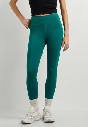Vrouw draagt teal high waist leggings, witte sokken, witte sneakers en een zwart crop top, staand met één hand op de heup.