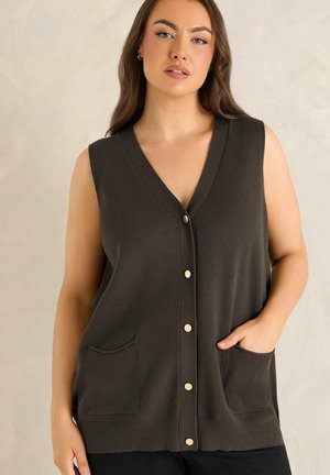 Femme portant un gilet sans manches marron foncé à boutons avec deux poches frontales, debout devant un fond neutre.