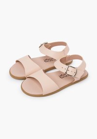 Vaaleanpunaiset nahkakengät, joissa on kaksi remmiä; toinen on solkikiinnityksellä ja toinen on leveä nauha. Litteä pohja, sileä pinta, minimalistinen muotoilu.