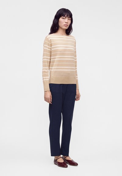 Femme aux longs cheveux noirs portant un pull beige rayé à manches longues, un pantalon bleu marine et des chaussures Mary Jane rouge foncé, debout devant un fond uni.