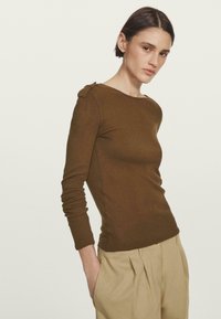 Brown long-sleeve top with a rounded neckline and side button detail, paired with beige pleated trousers. Smooth, fitted texture.