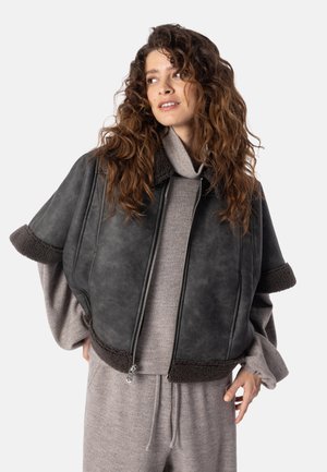 Frau mit langen lockigen Haaren trägt eine schwarze, mit Lammfell gefütterte kurze Jacke über einem grauen Strickrollkragenpullover und passenden Hosen.