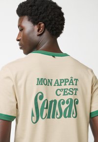 Beige T-shirt met korte mouwen en groene accenten. De achterkant heeft gedurfde witte tekst: "MON APPÂT C'EST SENSAS." Gladde textuur, casual ontwerp.