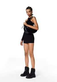 Sac en cuir noir porté à l'épaule avec un design courbé élégant, porté sur une robe noire côtelée. Le mannequin se tient dans des bottes noires chunky à lacets.
