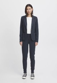 Navy-blau gestreifter Anzug mit einem maßgeschneiderten Blazer und eng anliegenden Hosen, kombiniert mit einem weißen Oberteil und schwarzen Sneakern. Weicher Stoff, strukturierte Gestaltung.