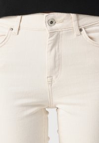 Jeans en denim blanc à coupe ajustée, dotés d'une fermeture par bouton métallique, de coutures discrètes et de poches avant. Texture lisse.