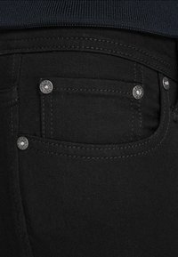 Los vaqueros de mezclilla negra presentan un primer plano de la zona del bolsillo, remaches metálicos plateados y costuras en contraste sobre una textura de tela suave.