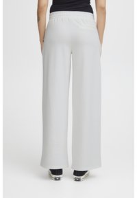 Helle graue weite Hose mit elastischem Bund und Gesäßtasche, kombiniert mit schwarzen Sneakern mit weißen Sohlen. Glatte Stoffstruktur.