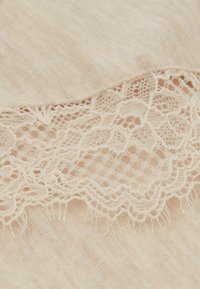 Beige tyg med en delikat spetskant längs kanten. Spetsen har blommönster och en vågig design, vilket skapar en kontrast i textur.