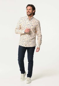 Overhemd met knopen in lichtgeel met een blauw bloemenpatroon, lange mouwen en een regular fit; gecombineerd met donkere jeans en witte sneakers.