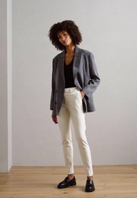 Blazer en daim gris sur un haut noir, associé à un pantalon blanc sur mesure et des mocassins noirs. Le modèle se tient devant un fond neutre.