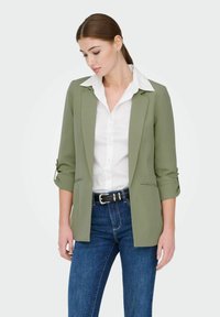 Blazer vert olive avec une coupe ajustée, doté d'un col cranté, de manches retroussées et de poches latérales, associé à une chemise blanche et un jean bleu.