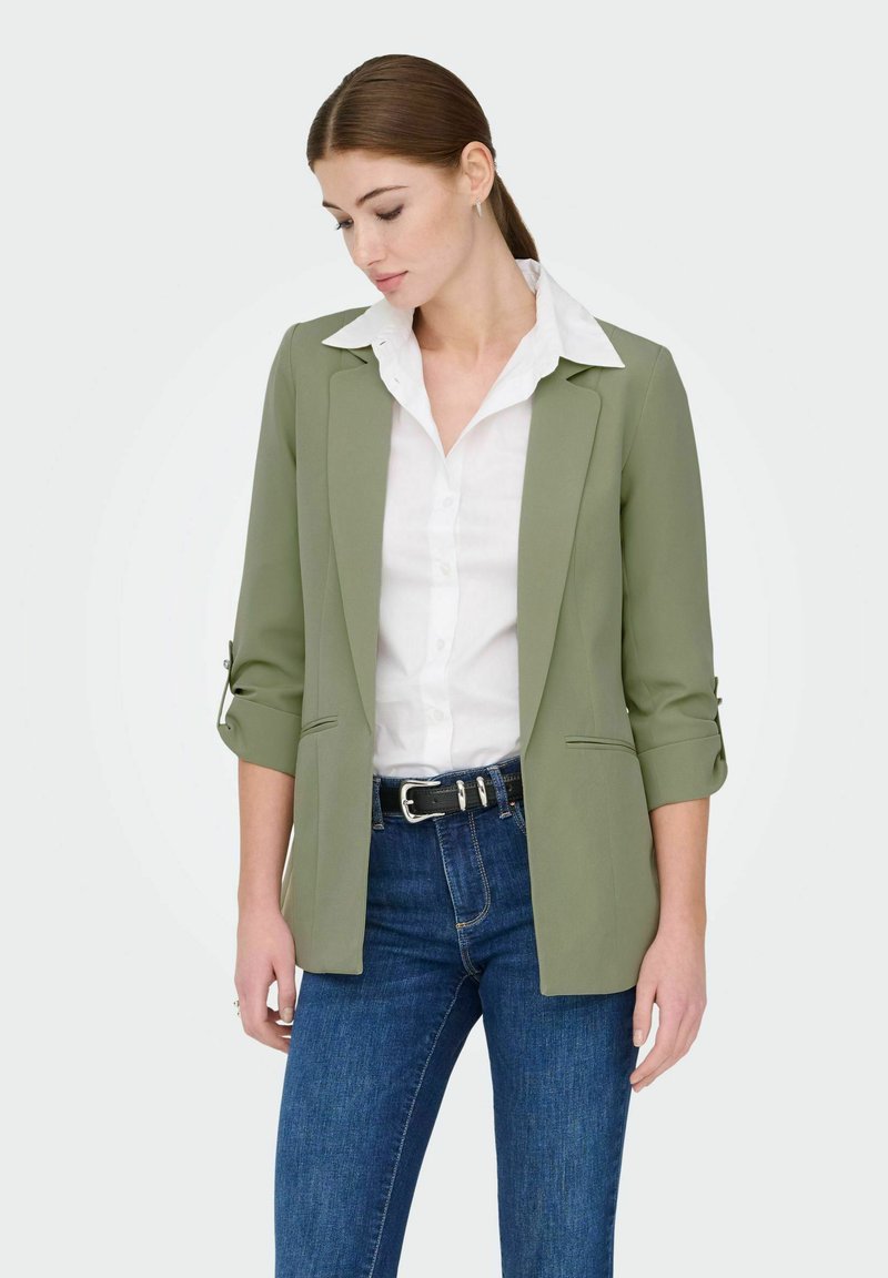 Blazer vert olive avec une coupe ajustée, doté d'un col cranté, de manches retroussées et de poches latérales, associé à une chemise blanche et un jean bleu.