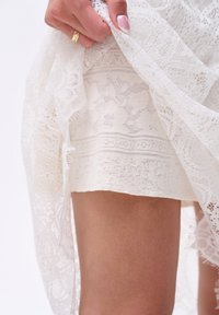 Robe en dentelle blanche présentant des motifs et textures complexes, superposée à une couche unie blanche lisse, avec un ourlet au milieu de la cuisse.