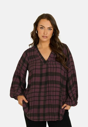 Vrouw draagt een losse paarse en zwarte geruite V-hals blouse met lange ballonmouwen, poseert met handen in de zakken tegen een effen achtergrond.