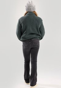 Faux fur green pullover, high collar, black flared jeans, casual white-soled shoes, woman wearing a striped beanie, viewed from the back.