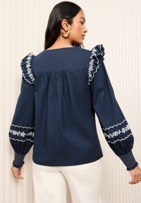 Navy blue long-sleeve blouse with ruffled shoulders, floral embroidery, and striped cuffs. Soft fabric with subtle pleating at the back.