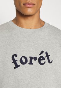 Close-up van een persoon die een lichtgrijze sweatshirt draagt met het woord "forét" in donkerblauw geborduurd op de borst.