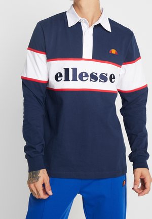 Homme portant un polo à manches longues bleu marine, blanc et rouge avec le logo "ellesse" et un pantalon bleu vif, avec un tatouage visible sur la main gauche.