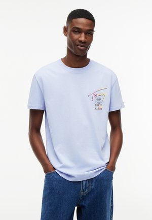Junger Mann trägt ein hellviolettes T-Shirt mit Grafik und blaue Jeans, steht mit Händen in den Taschen vor weißem einfarbigem Hintergrund.