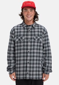 Szara i czarna koszula flanelowa w kratę z długimi rękawami, zapinana na guziki, z dwoma kieszeniami na piersi i teksturowaną tkaniną. Noszona z czerwoną czapką.