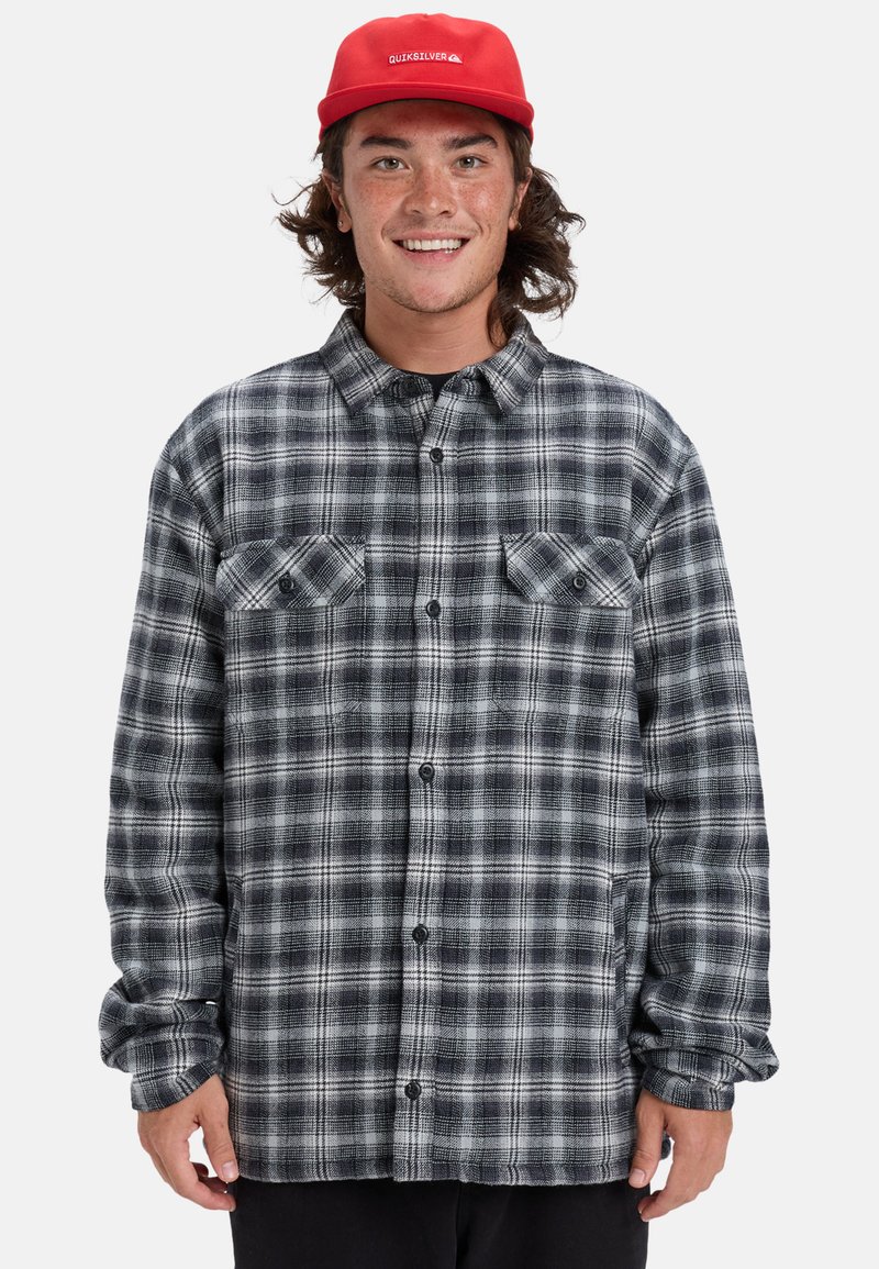 Szara i czarna koszula flanelowa w kratę z długimi rękawami, zapinana na guziki, z dwoma kieszeniami na piersi i teksturowaną tkaniną. Noszona z czerwoną czapką.