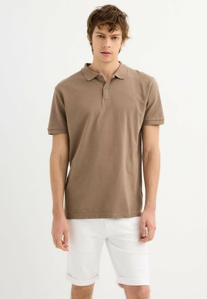 Camisa polo de algodón marrón con cuello clásico, mangas cortas y un pequeño logo en el pecho, combinada con pantalones cortos blancos con los puños doblados.