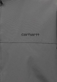 Γκρίζο τζάκετ με υφασμάτινη επιφάνεια που είναι ανθεκτική στο νερό. Διαθέτει ραμμένο τσέπη και μαύρο κεντημένο λογότυπο "carhartt".