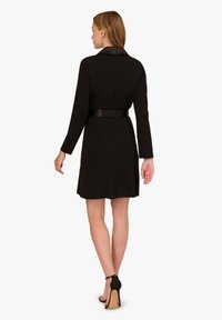 Robe noire à manches longues et à taille ceinturée. Dotée d'un col classique et d'un ourlet mi-cuisse. Texture de tissu lisse.