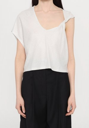 Kvinde iført en hvid asymmetrisk top med korte ærmer og en knude-detalje på skulderen samt sorte højttaljede bukser.