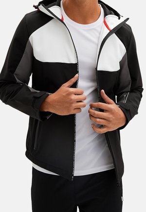 Person iført en sort og hvid lynlåssweatshirt over en hvid skjorte, med hænder der justerer lynlåsen ved brysthøjde.